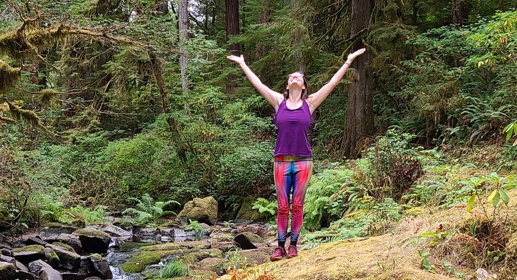 Sara, white woman in rainbow leggings arms raised, looking up, in the forest next to a stream