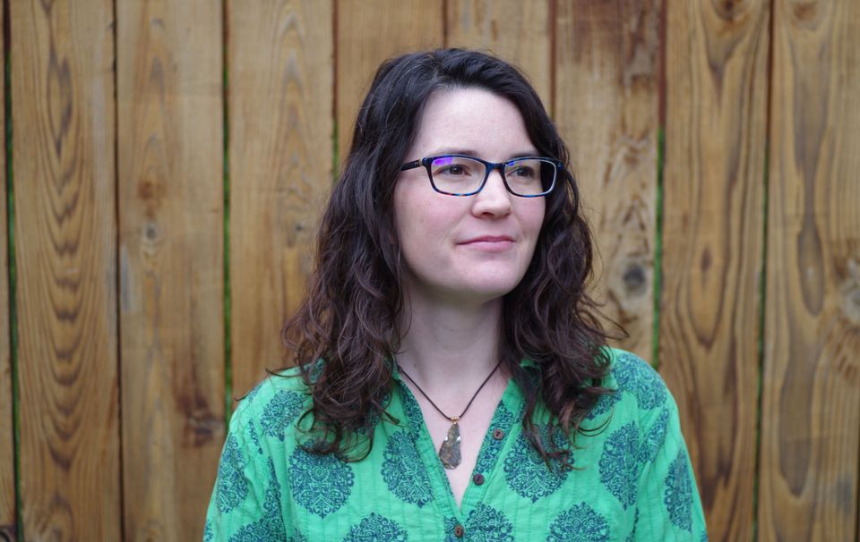 Photo of Sara Clark looking into the distance, slight smile. Bright green shirt, brown hair down, white woman wearing glasses