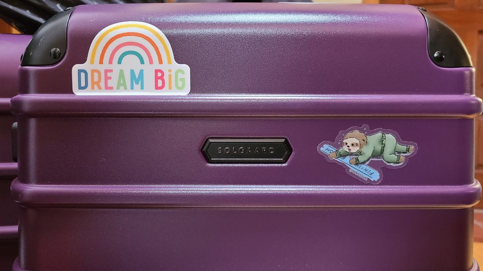 new purple suitcase with stickers "dream big" and an exhausted sloth crossing a finish line