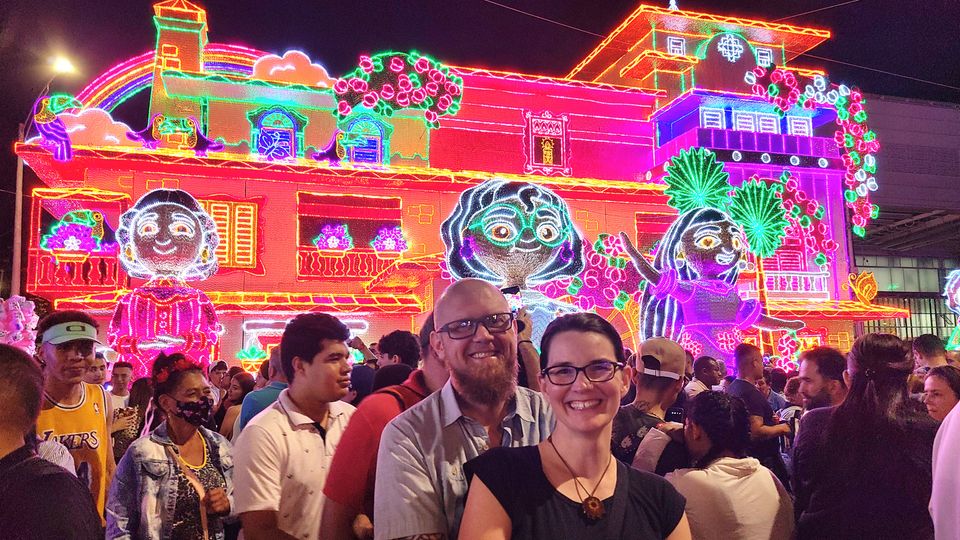 Brian and Sara smiling, lots of people walking around behind them and a lit up model of the house in the Encanto movie.